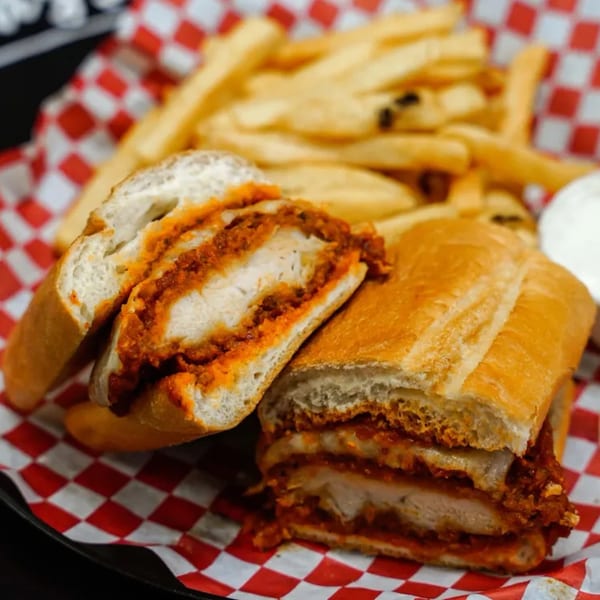 Chicken parmesan hero sandwich with fries and dipping sauce on a red and white checkered paper.
