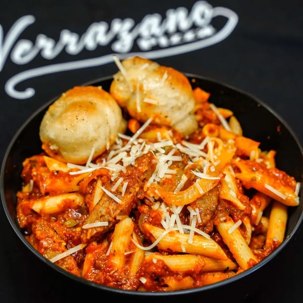 Penne pasta with red sauce, sausage, onions, shredded cheese, and two garlic knots in a black bowl.