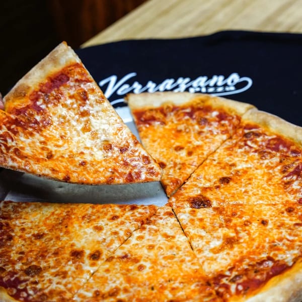 A hand lifts a slice of Verrazano Pizza's classic cheese pizza from a whole pie.