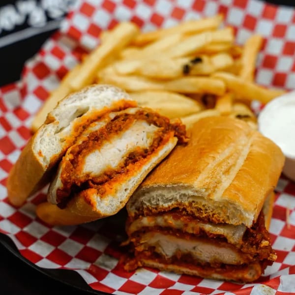 Chicken parmesan hero sandwich with marinara sauce and fries on a red and white checkered paper.